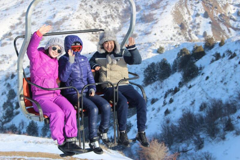 Three people are riding a ski lift, waving, with a snowy mountain in the background.