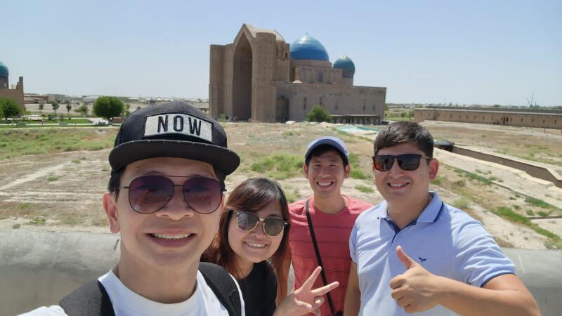 A selfie of four people wearing sunglasses, with a building with blue domes in the background. One person wears a cap with the word "NOW" on it.