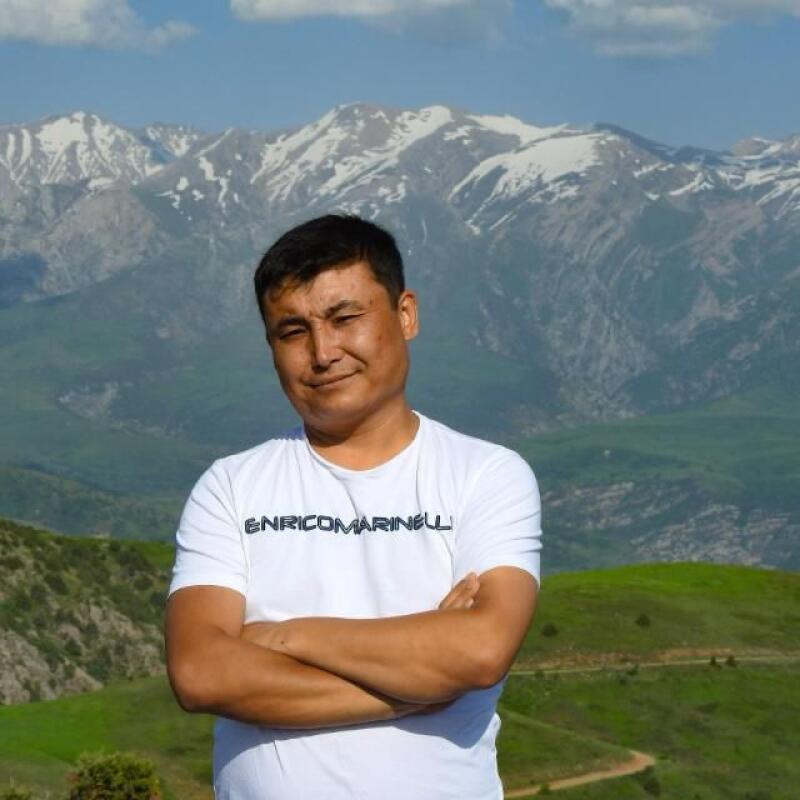 A man with his arms crossed is wearing a white T-shirt with "ENRICOMARINELLI" printed on it, with a mountain range in the background.