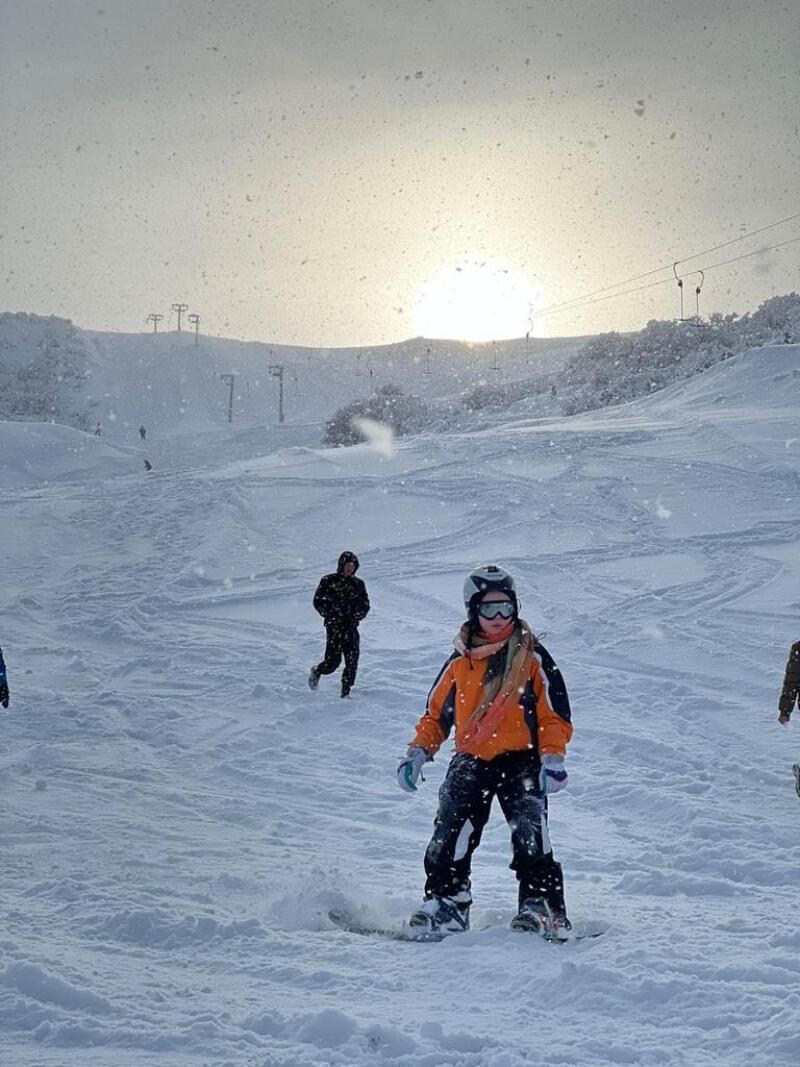 A snowboarder in an orange and black jacket stands in the snow, with other people and ski lifts visible in the snowy, mountainous background.