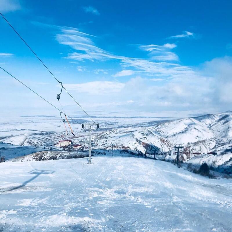 A snowy mountain landscape with a ski lift under a blue sky with white clouds.