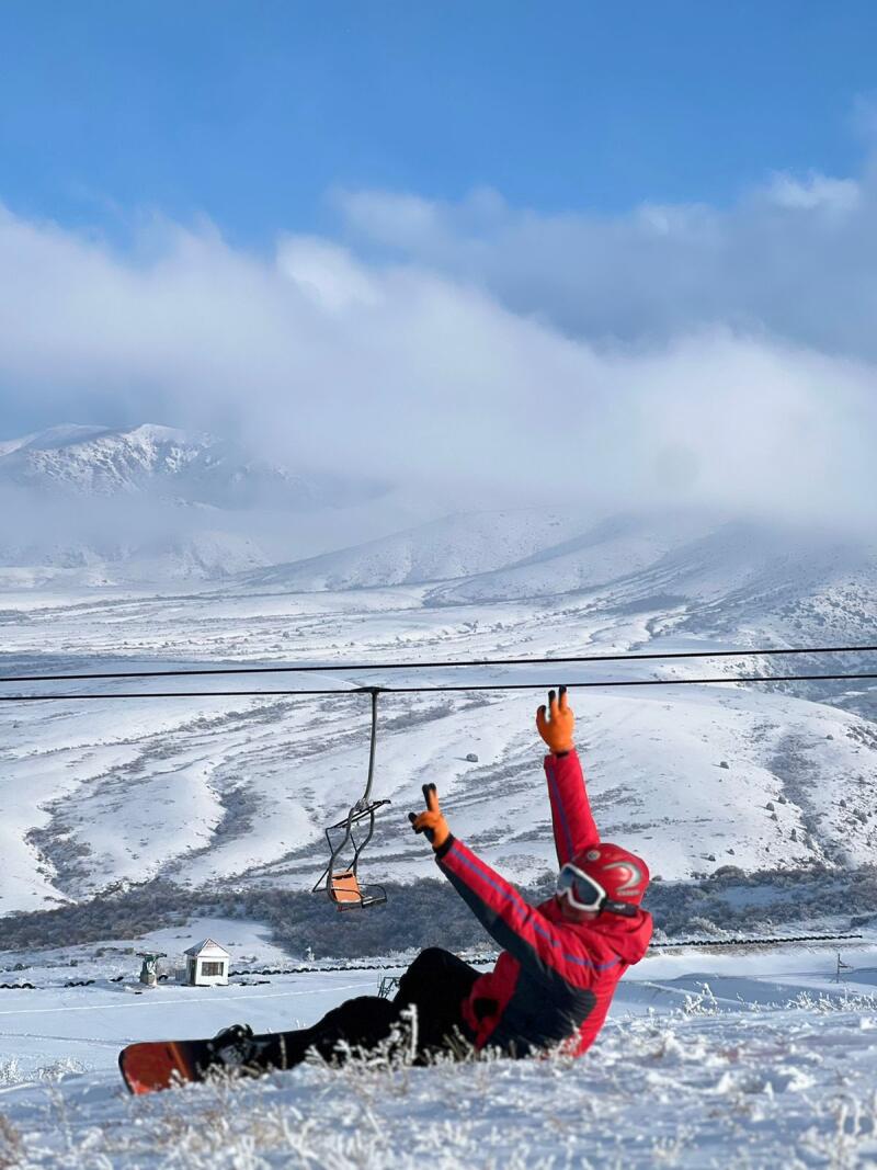 A snowboarder in a red jacket, helmet, and goggles sits in the snow with arms raised in a V shape, in front of a snowy mountain landscape and a chairlift.