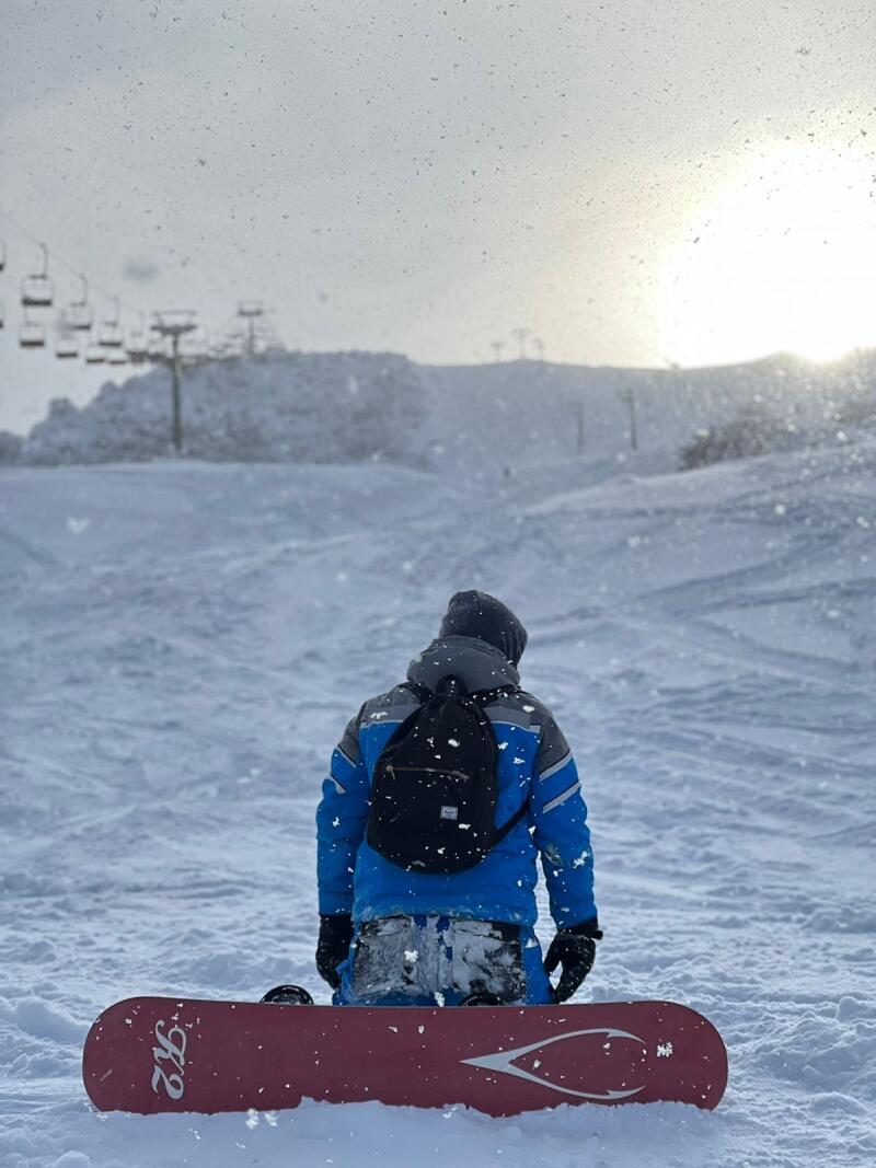 A person in a blue jacket, black gloves, and a gray hat is kneeling in the snow with a red snowboard, looking at a ski slope with a ski lift on a snowy day.