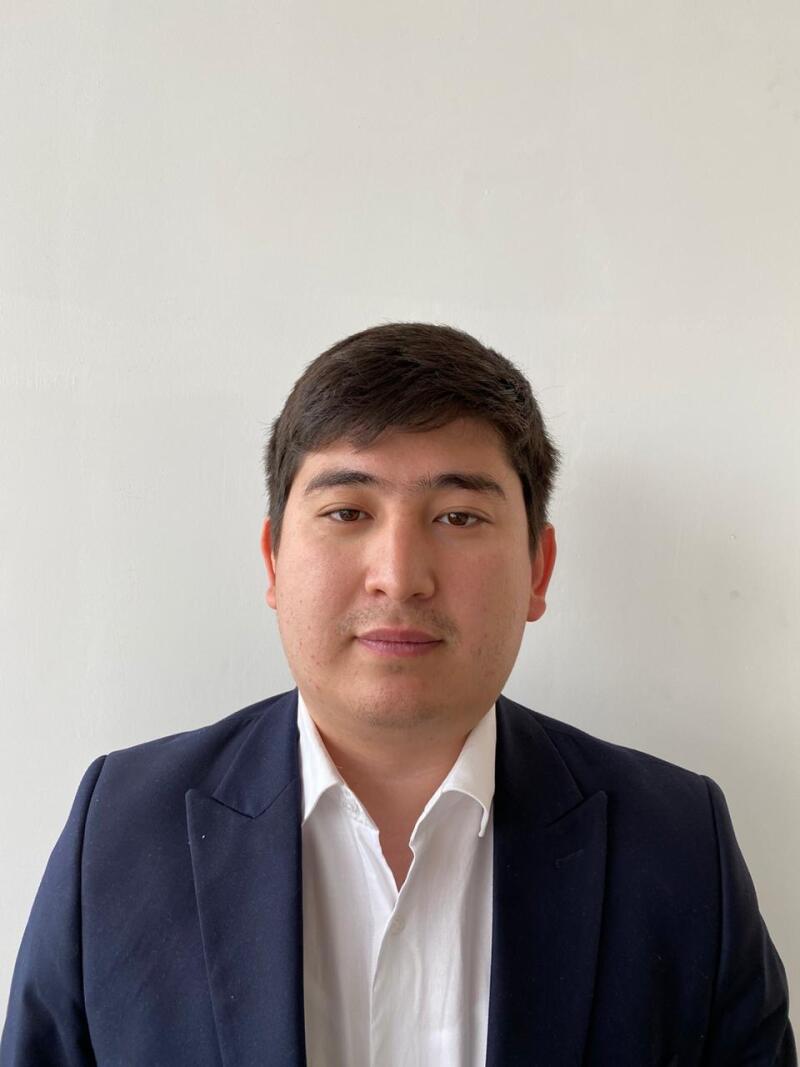 A man in a white collared shirt and a navy blue blazer stands against a white background.