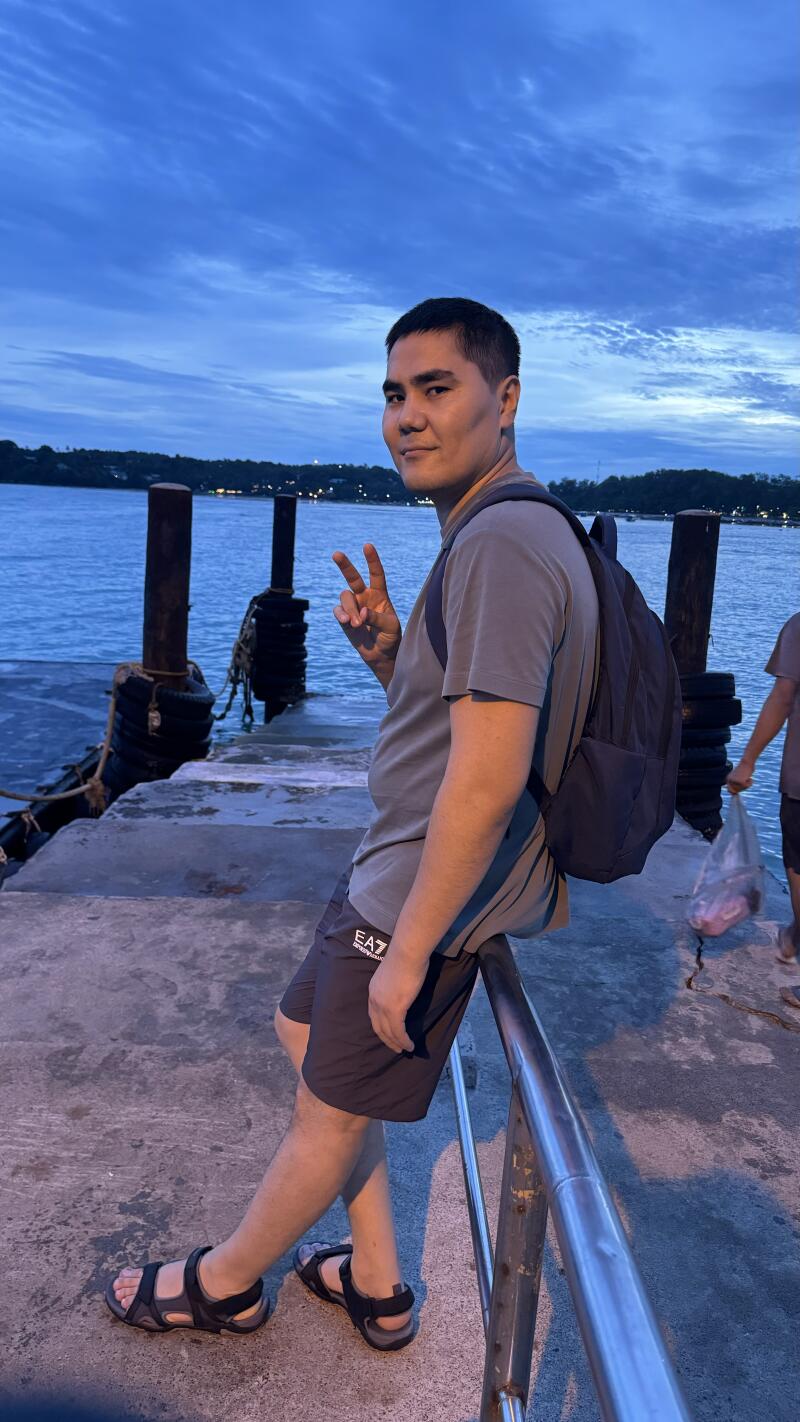 A man in a gray shirt and shorts with "EA7" printed on them is sitting on a railing with a backpack and making a peace sign in front of a body of water.