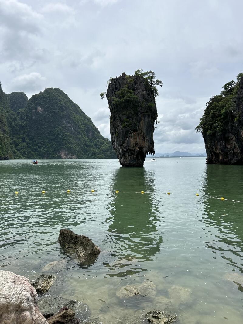 The image shows a tall, rocky island with some greenery on top, standing in a body of water. There are mountains in the background and some yellow buoys floating on the surface of the water.