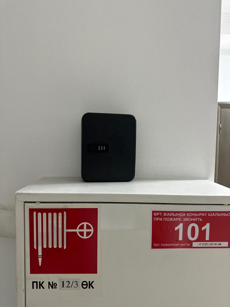 A black combination lock box is mounted above a white fire hose cabinet with red signage, including instructions to call '101' in case of fire and the number '+7 (727) 337-91-98'.