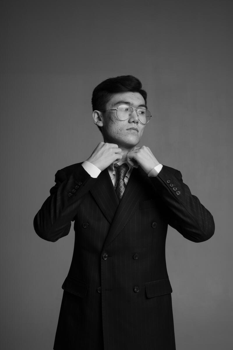 A man in a pinstripe suit and glasses adjusts his collar in a black and white portrait.
