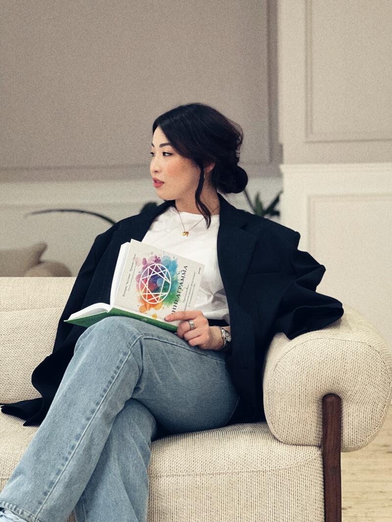 A woman with dark hair sits on a light-colored couch, wearing a black blazer and blue jeans, and holds a book titled "ЭННЕАГРАММА" with a colorful geometric design on the cover.