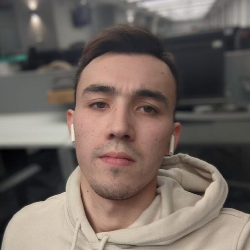 A selfie of a man with short dark hair wearing a beige hoodie and white wireless earbuds, set against an office background.