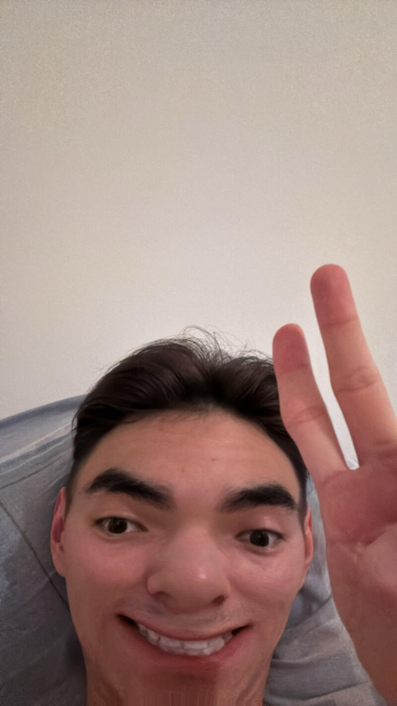 A selfie of a smiling person with dark hair, holding up two fingers in a peace sign.
