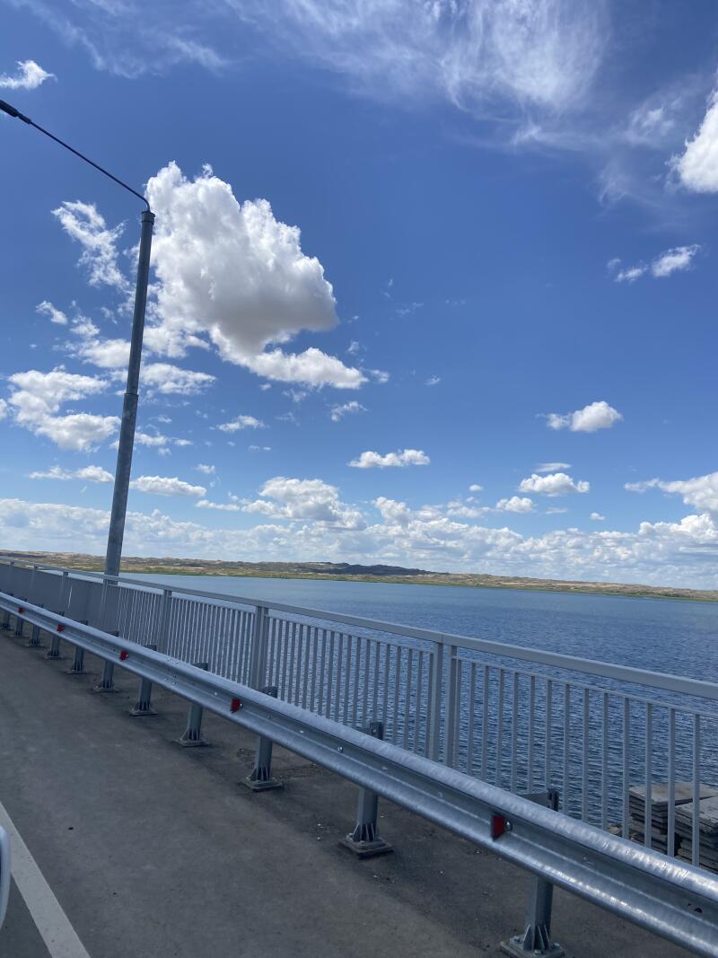 Вид с моста на большое синее водное пространство с далекой сушей, под ярким голубым небом с белыми облаками. С левой стороны моста виден фонарный столб и металлический отбойник.