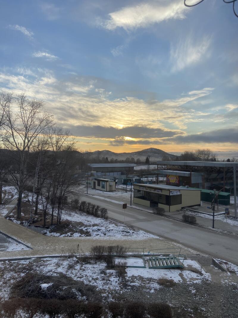 Зимний пейзаж на восходе или закате, показывающий заснеженную землю, голые деревья и дорогу. Небольшие здания с видимыми вывесками, такими как «БАРЫС» и «САУНА-АУЫЛ», расположены вдоль дороги, с горами и большой крытой конструкцией на заднем плане под облачным небом с золотистым светом.