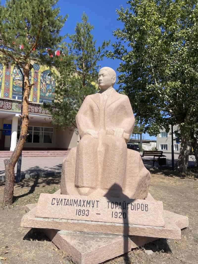 Светло-коричневая каменная статуя сидящего человека, Султанмахмута Торайгырова, с его именем и годами жизни «1893 - 1920», высеченными на основании, стоит на открытом воздухе перед зданием с красочной мозаикой и деревьями под голубым небом.