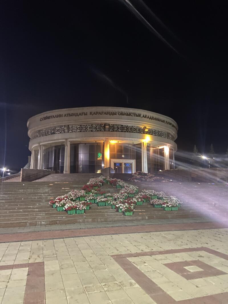 Ярко освещенное круглое здание ночью, с парадной лестницей, украшенной многочисленными горшечными красными и белыми цветами. На фасаде здания виден текст: «СЕЙФУЛЛИН АТЫНДАҒЫ ҚАРАҒАНДЫ ОБЛЫСТЫҚ АКАДЕМИЯЛЫҚ».
