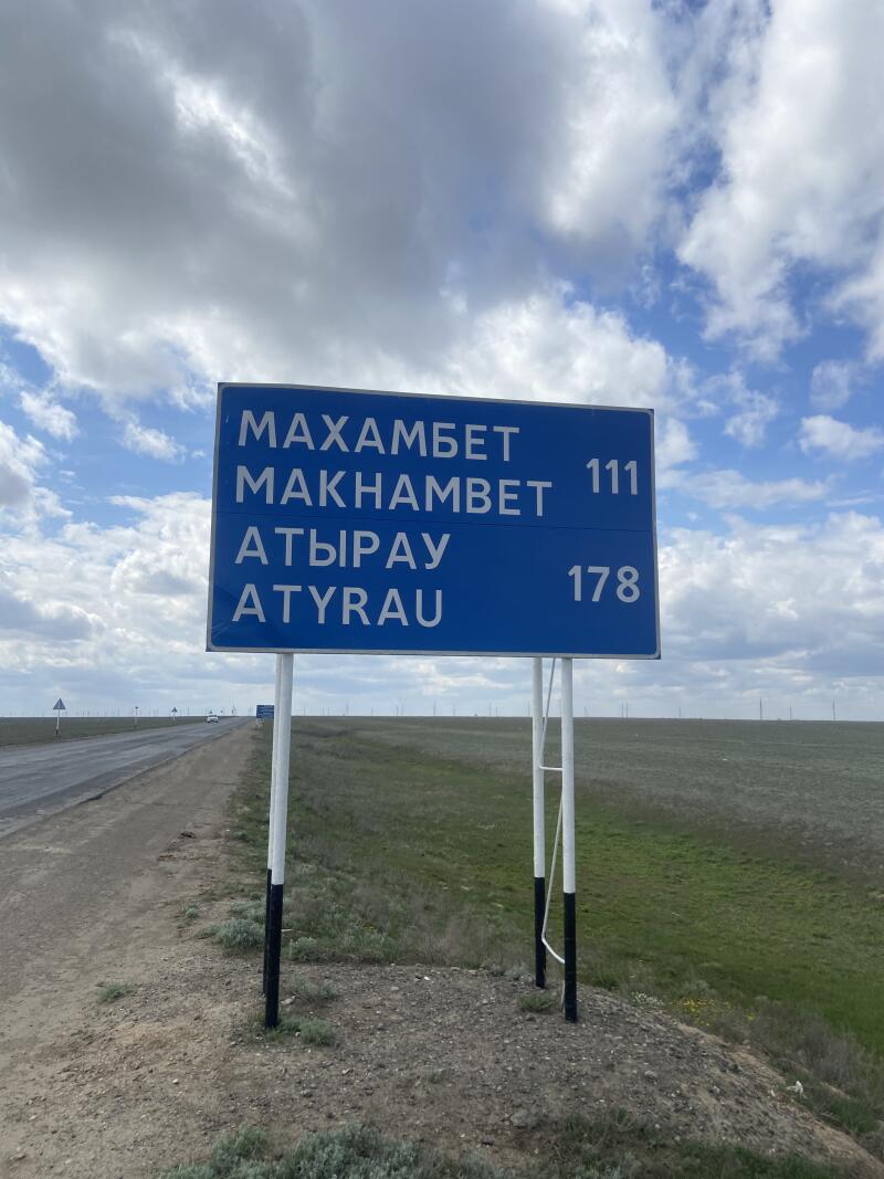 Синий дорожный знак с белым текстом стоит у дороги с зелеными полями под облачным небом. На знаке написано «МАХАМБЕТ», «МАКНАМБЕТ 111», «АТЫРАУ» и «ATYRAU 178».