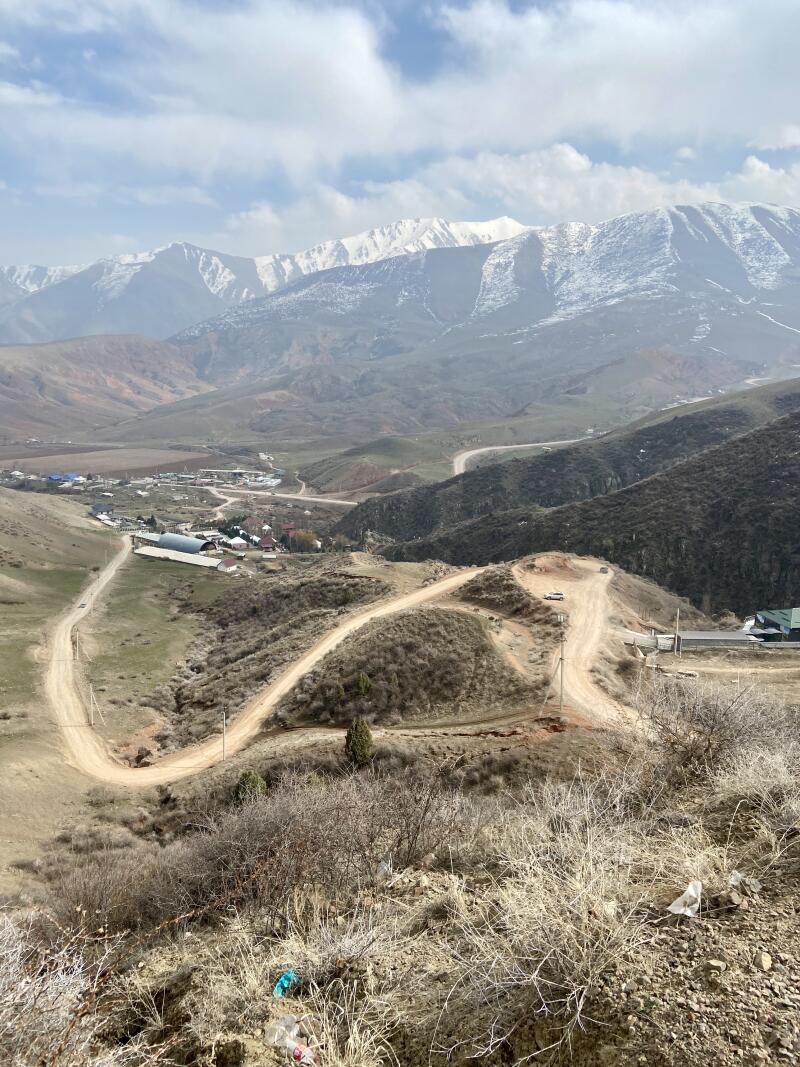 Панорамный вид на долину с деревней и извилистыми грунтовыми дорогами, ведущими к заснеженным горам под облачным небом, с сухой растительностью и мусором на переднем плане.