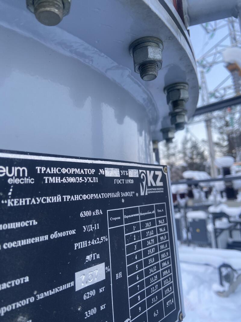 Крупный план серого промышленного трансформатора с черной табличкой, на которой указаны характеристики, такие как "ТРАНСФОРМАТОР ТМН-6300/35-УХЛ1", "КЕНТАУСКИЙ ТРАНСФОРМАТОРНЫЙ ЗАВОД", "6300 кВА", и таблица значений напряжения и тока. На размытом заднем плане виден снег на земле.