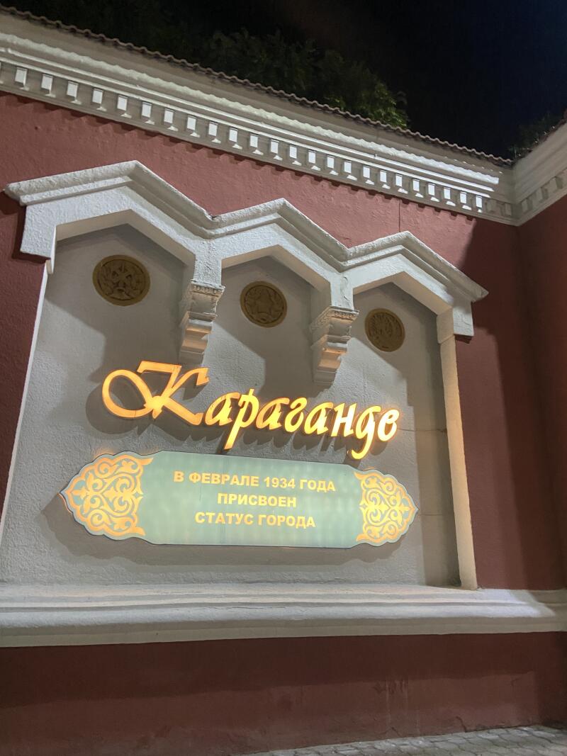 Светящаяся вывеска "Караганды" размещена на фасаде здания с табличкой внизу, на которой написано "В феврале 1934 года присвоен статус города". Фасад украшен белым декоративным молдингом и золотыми медальонами.