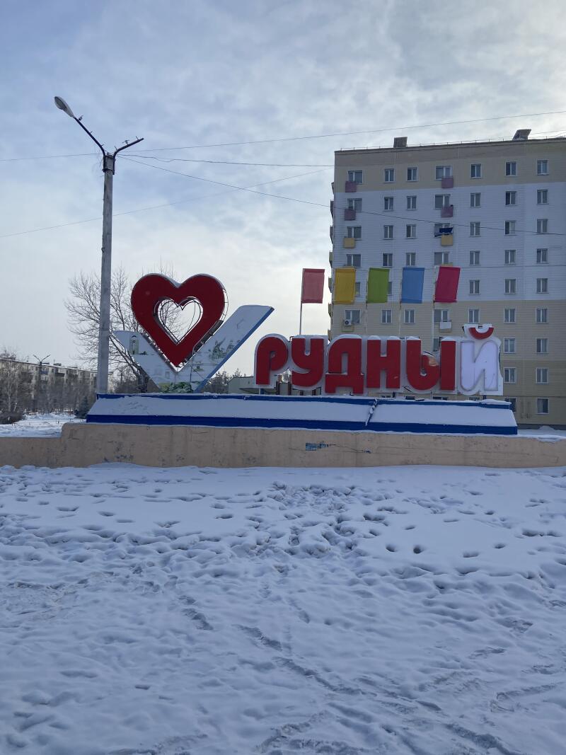 Занесенная снегом приветственная стела с большим красным сердцем и красным текстом «РУДНЫЙ» установлена на постаменте посреди заснеженного пейзажа. На заднем плане, под пасмурным небом, виднеется многоэтажное здание с разноцветными флагами.