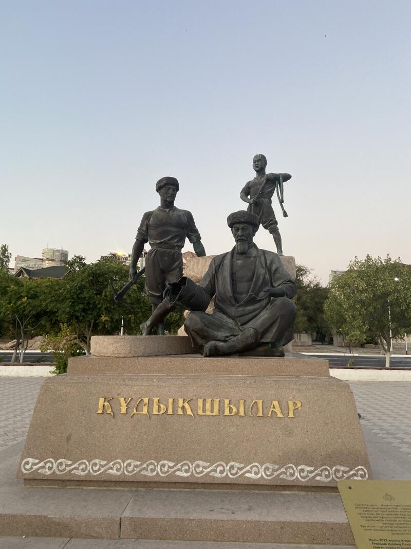 Бронзовый памятник изображает трех фигур: мужчину, сидящего со скрещенными ногами на переднем плане, мужчину, стоящего слева с инструментом, и мужчину, стоящего справа с веревкой. Памятник установлен на каменном постаменте с позолоченной надписью на казахском языке "ҚҰДЫҚШЫЛАР".