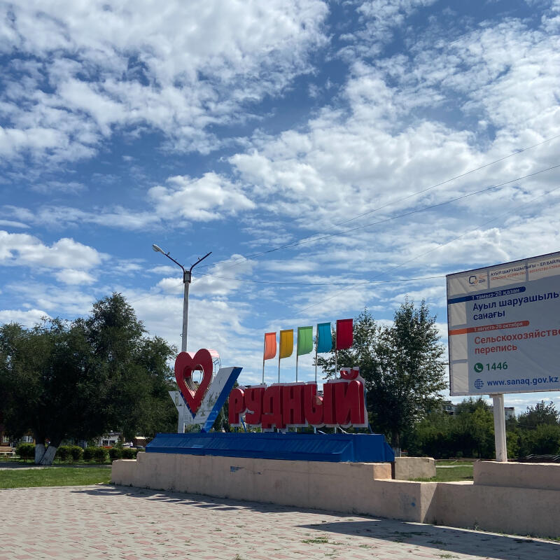 Большая надпись «Я ❤️ РУДНЫЙ» с красными буквами и сердцем расположена в парковой зоне с зелеными деревьями и травой под облачным небом. Справа от нее видны разноцветные флаги и рекламный щит, анонсирующий сельскохозяйственную перепись с текстом «АУЫЛ ШАРУАШЫЛЫҒЫ санағы» и «Сельскохозяйственная перепись», датами, номером телефона «1446» и сайтом «www.sanaq.gov.kz».
