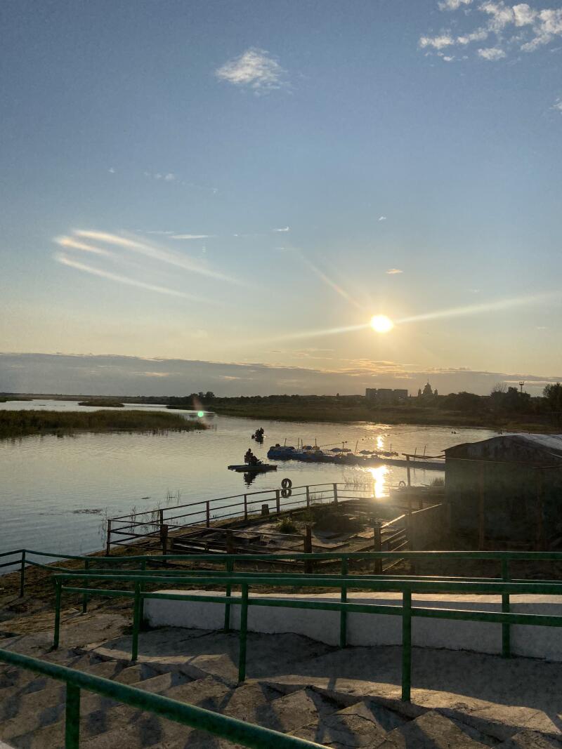 Вид на закат над широкой рекой с низким солнцем, отражающимся в воде, лодками с людьми и бетонной лестницей с зелеными перилами на переднем плане. На горизонте видны здания.