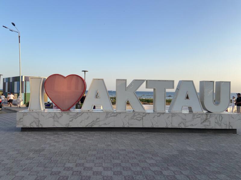 Большой белый знак «I ❤️ AKTAU» с красным сердцем, на постаменте с мраморным узором, стоит на плиточной мостовой на фоне моря и чистого голубого неба.