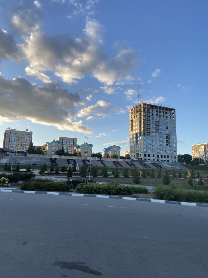 Городской пейзаж с высоким строящимся зданием и краном, несколькими жилыми зданиями с синими крышами и большой надписью «ҚОСТАНАЙ» на насыпи перед ними, все под частично облачным небом.