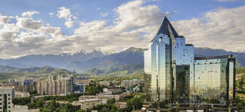 The image shows a cityscape featuring modern buildings and a glass-clad skyscraper with a pyramid-shaped top, set against a backdrop of mountains and a cloudy sky.