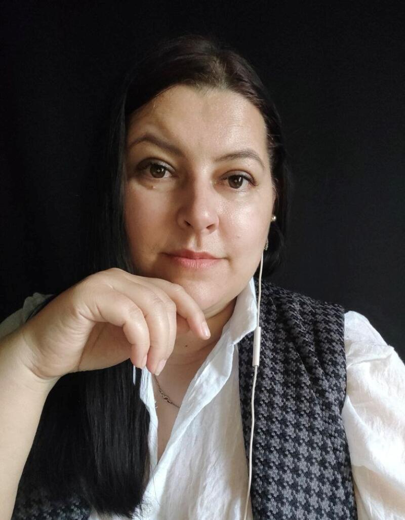 A woman with dark hair is looking at the camera with her hand near her face, wearing a white shirt and a patterned vest against a dark background; she also has white earphones in her ears.