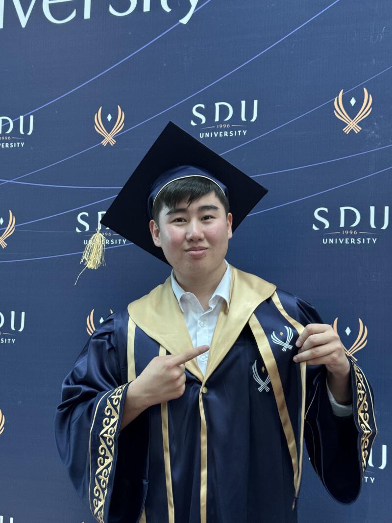A graduate in a dark blue gown and cap stands against a backdrop featuring the SDU University logo, pointing at it with his right hand.