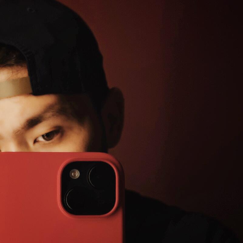 A person wearing a black cap is partially visible behind a red smartphone, with only their eye and part of their face showing.