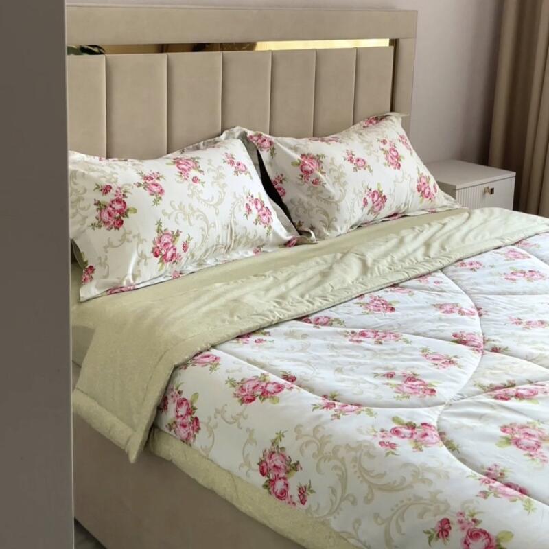 A bed is neatly made with floral-patterned bedding in shades of white and pink, complemented by a light green duvet and pillowcases, all set against a beige headboard and a matching bedside table.