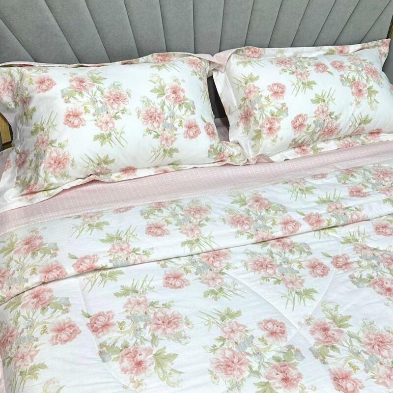 A bed with a gray headboard is covered in white bedding with a floral pattern of pink flowers and green leaves. Two pillows with the same floral pattern are placed at the head of the bed, and a light pink sheet is visible between the pillows and the comforter.