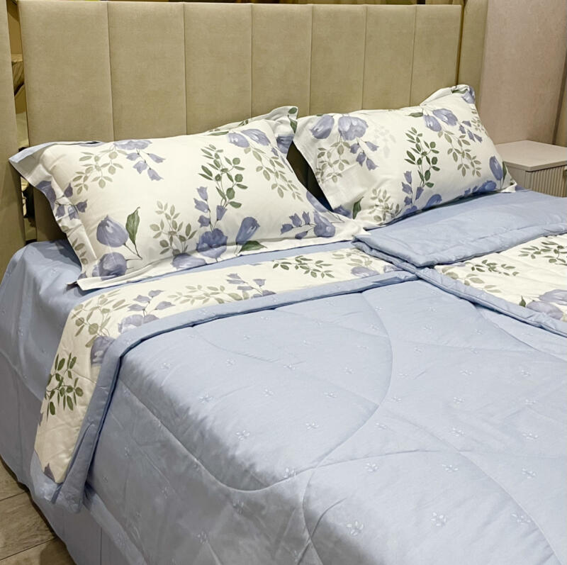 The image shows a made bed with light blue sheets and a floral-patterned comforter and pillows. The headboard is a neutral beige color, and a nightstand is visible in the background.