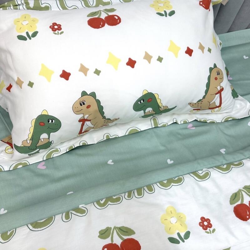 The image shows a bed with a pillow and bedding set featuring cartoon dinosaurs, flowers, cherries, and star-shaped patterns on a white background with light green accents.