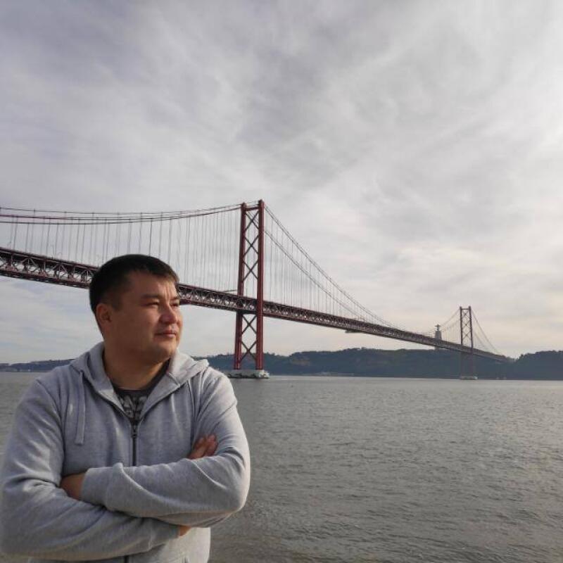 A man with folded arms is standing in front of a body of water and a red suspension bridge under a cloudy sky.