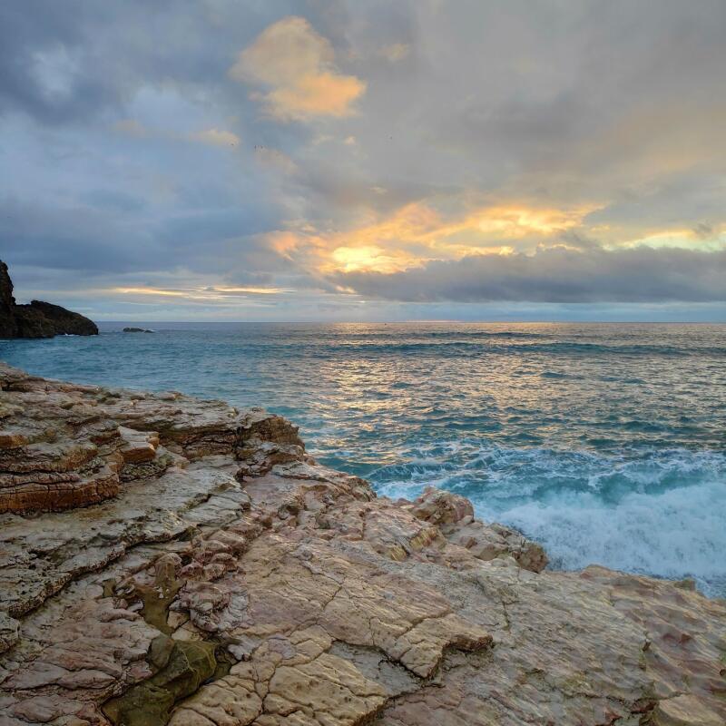 A scenic view of a rocky shoreline meeting a wavy ocean under a cloudy sky at sunset.