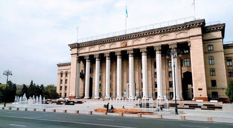 The image shows a large, light-colored building with many columns, likely a government or civic building, with a flagpole on top and a fountain in the foreground.