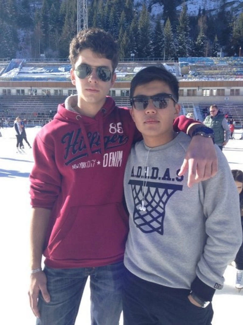 Two young men wearing sunglasses and casual clothing stand close together in front of an outdoor ice rink and a backdrop of pine trees and stadium seating.