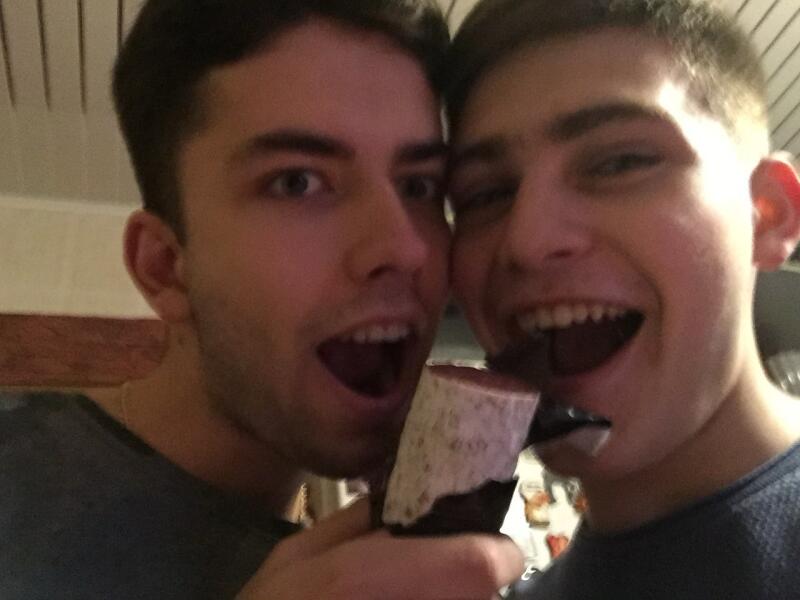 Two men with open mouths are about to take a bite of a dessert with chocolate and white cream.
