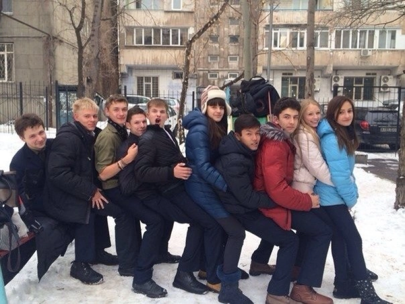 A group of about ten young people, mostly teenagers, are standing in a line outside in winter, each sitting on the lap of the person behind them.