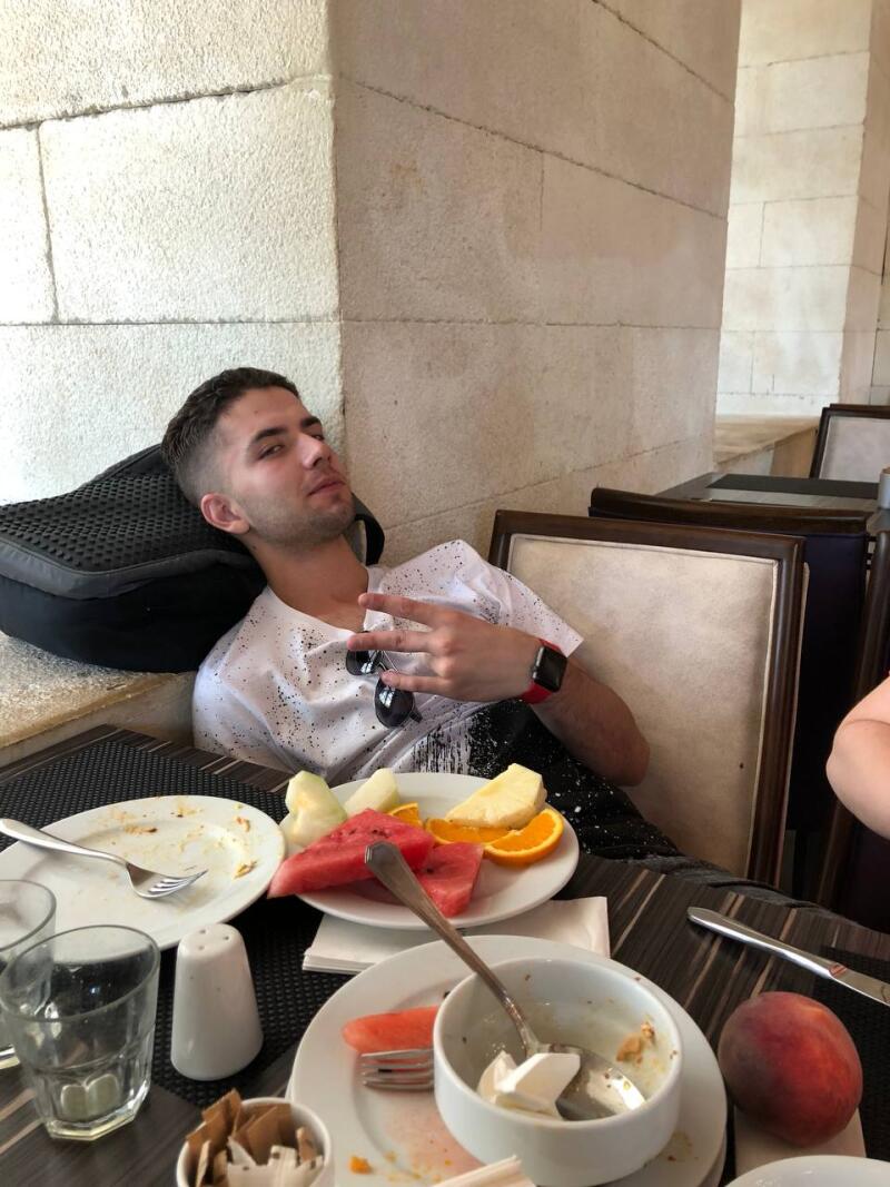A young man is sitting at a table with plates of fruit, gesturing with his hand while wearing sunglasses and a smartwatch.