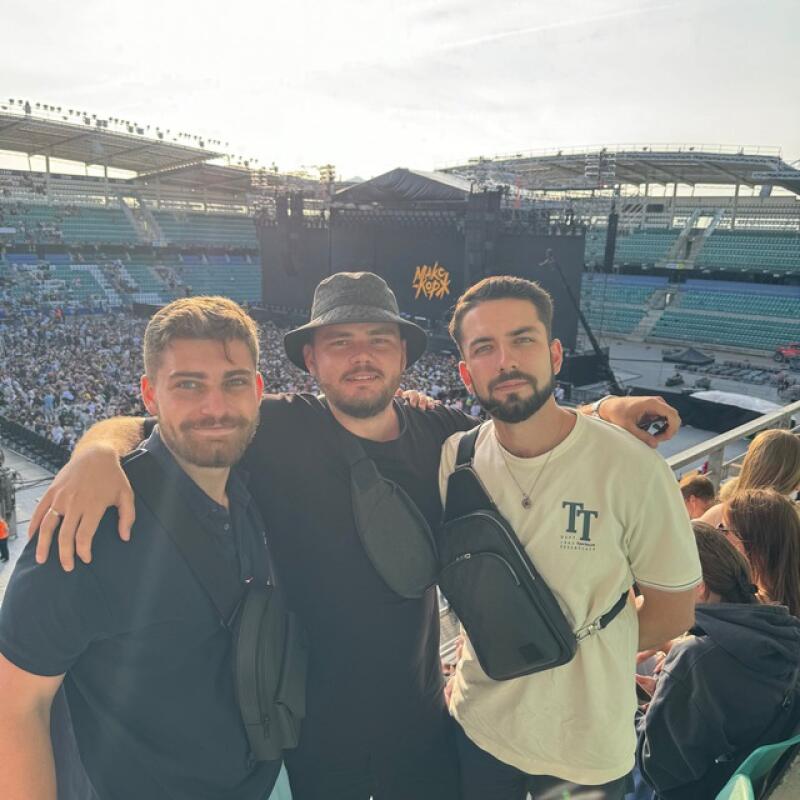 Three men are posing in front of a stage at an outdoor concert; the backdrop features the words "MIKEY KOPЖ".