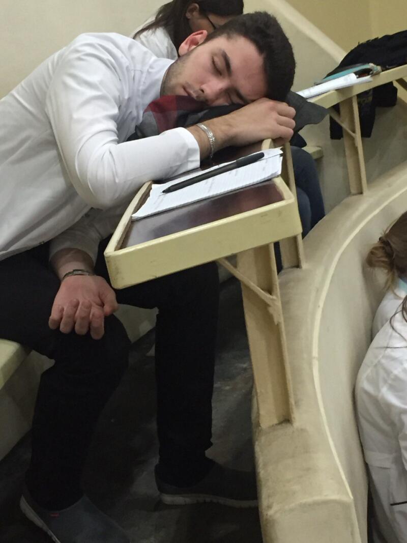 A man in a white shirt is asleep with his head on a desk in a lecture hall.