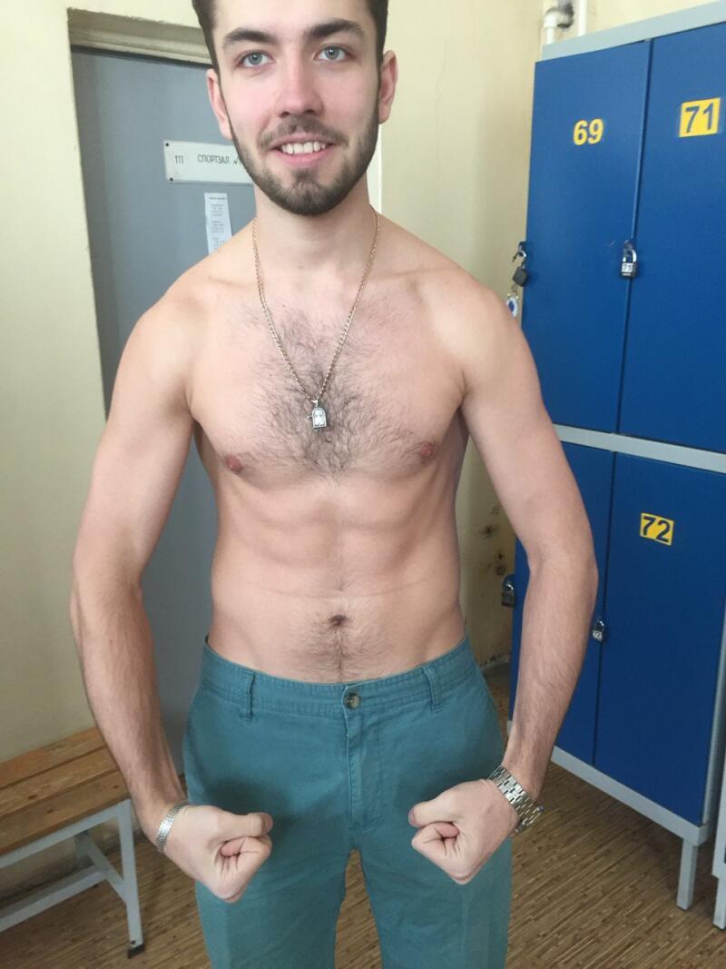 A man with a bare torso, wearing a necklace and turquoise pants, is flexing his arms in front of blue lockers numbered 69, 71, and 72.