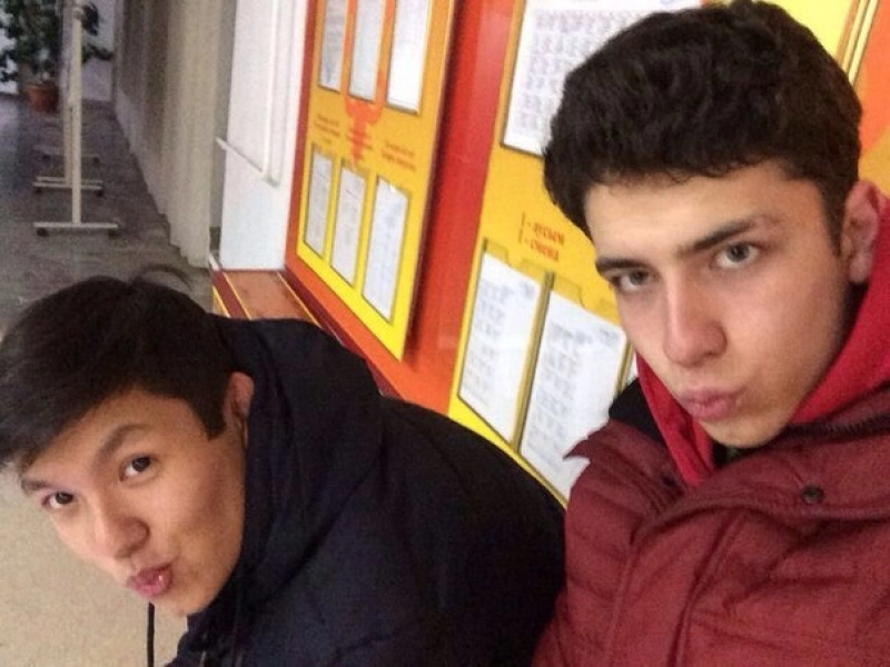 Two young men are posing for a selfie with puckered lips, wearing dark jackets. Behind them is a yellow wall with framed papers containing text.