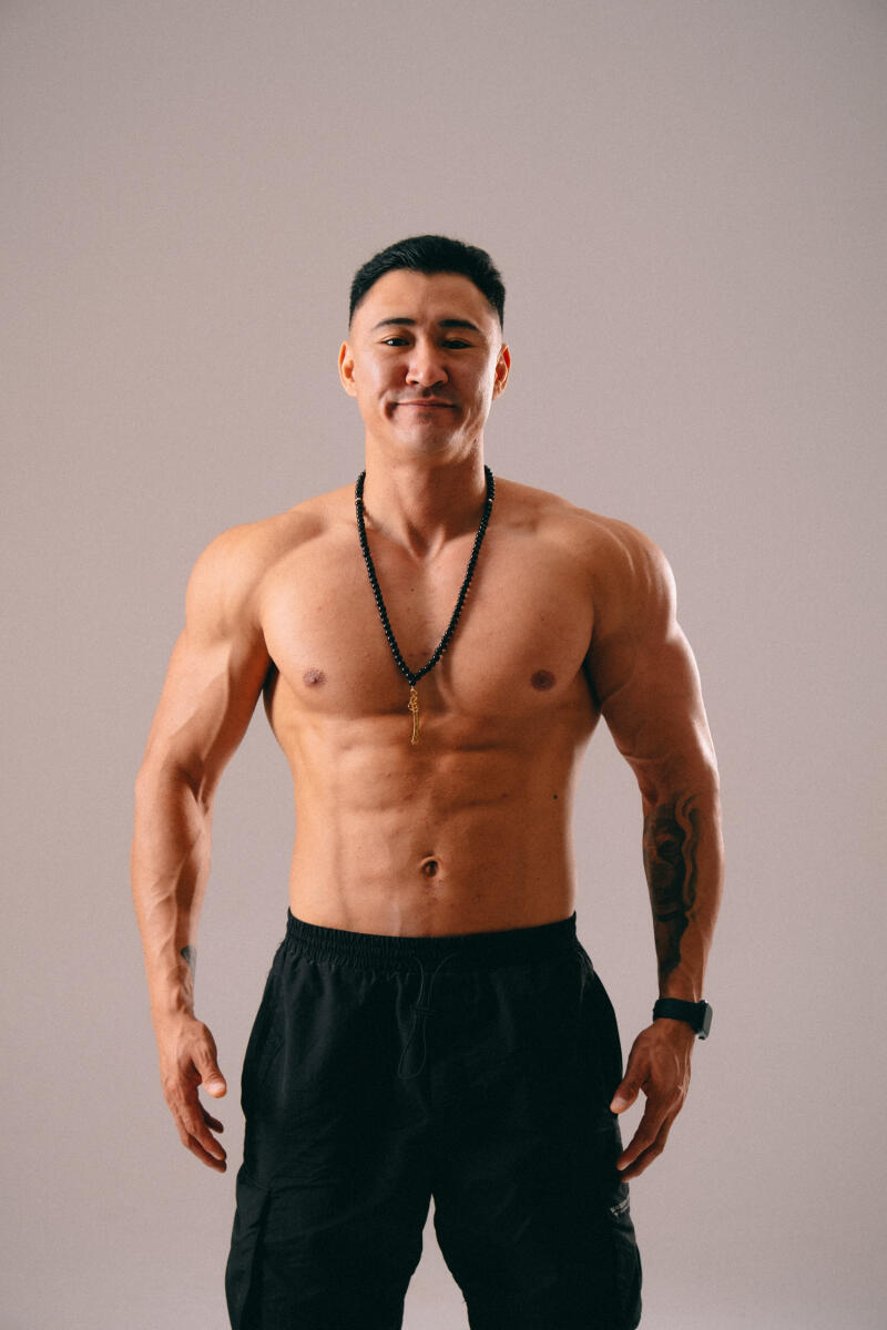 A muscular man with a beaded necklace stands shirtless in black shorts, displaying tattoos on his arms against a neutral background.