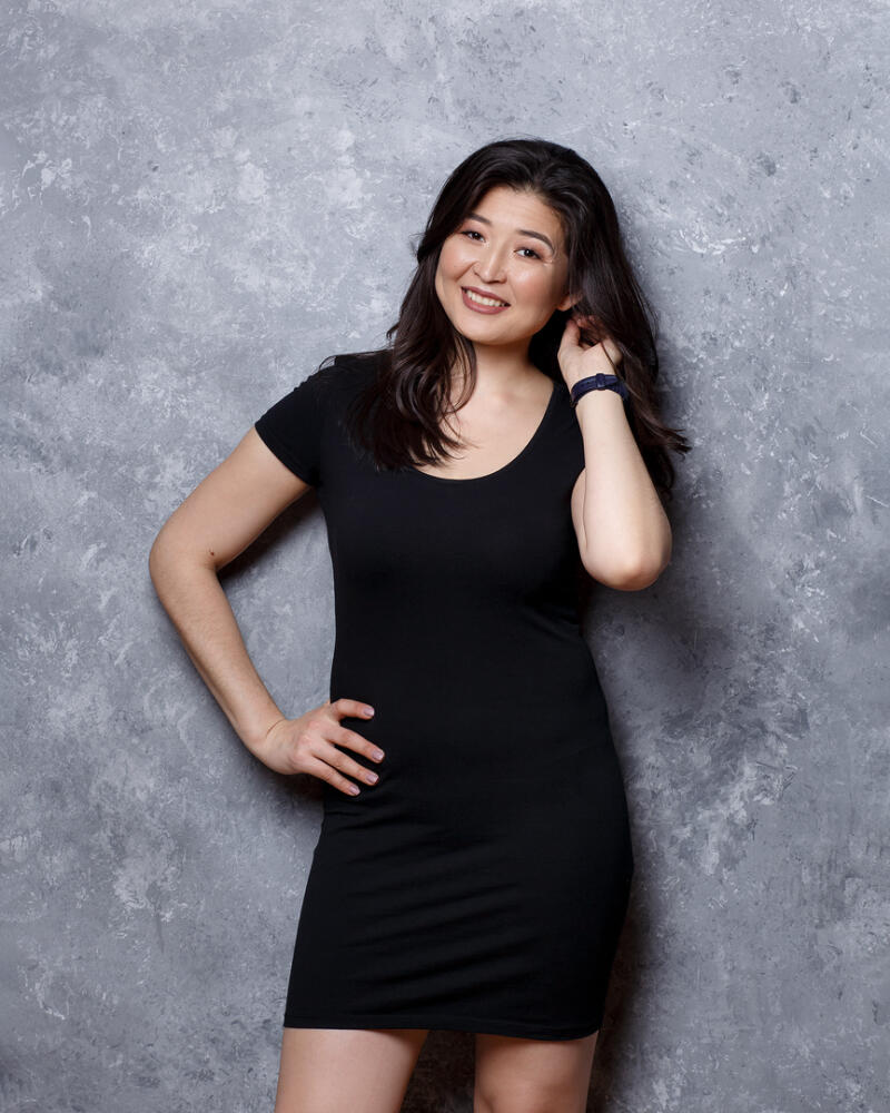 A smiling woman with dark hair stands against a gray mottled background, wearing a black dress and a dark wristband.